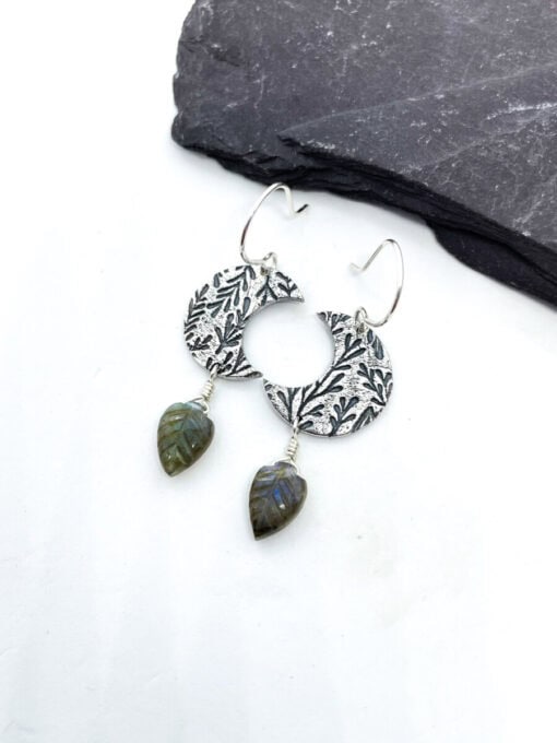 Silver earrings with circular textured design and leaf-shaped green stone pendants, placed on a gray slate slab.