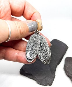 Hand holding silver earrings shaped like textured leaves with crescent moon designs, against a white background with dark stones.