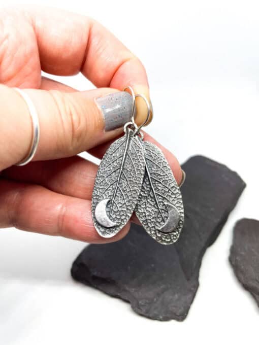 Hand holding silver earrings shaped like textured leaves with crescent moon designs, against a white background with dark stones.