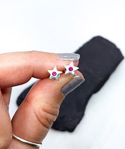 Hand holding small star-shaped silver earrings with pink gemstones in the center, against a white background.