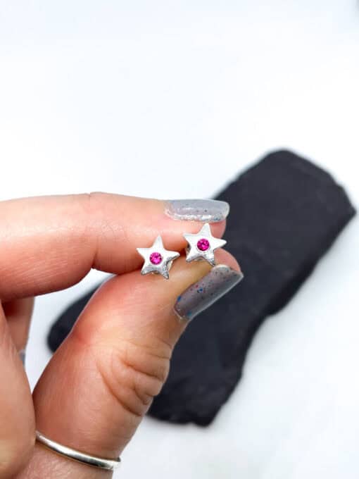 Hand holding small star-shaped silver earrings with pink gemstones in the center, against a white background.