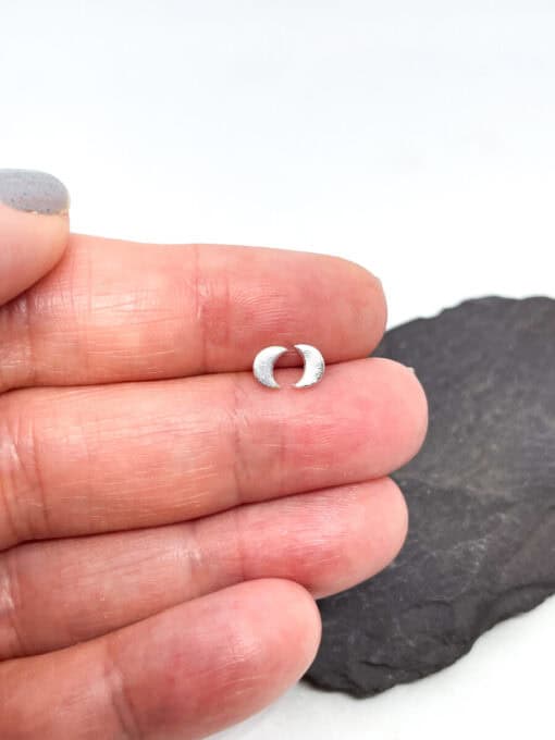 Close-up of a hand holding a small crescent moon-shaped silver stud earring, with a dark rock background.