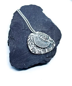 Silver etched necklace with a layered leaf and crescent moon design, displayed on a dark slate stone on a white background.