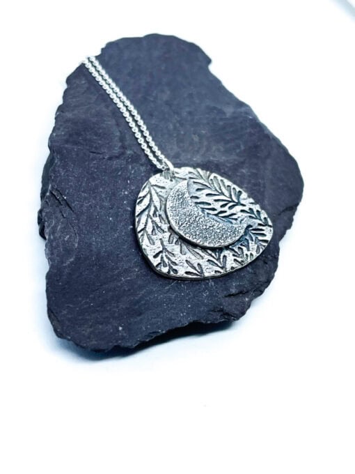 Silver etched necklace with a layered leaf and crescent moon design, displayed on a dark slate stone on a white background.