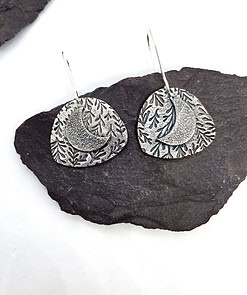 Silver crescent moon earrings with leaf patterns on a dark slate background.