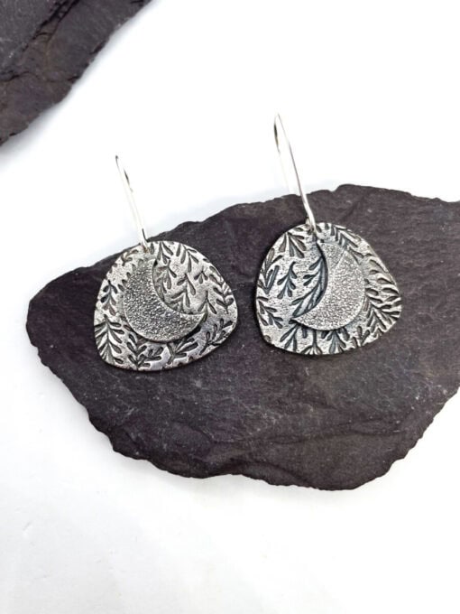 Silver crescent moon earrings with leaf patterns on a dark slate background.