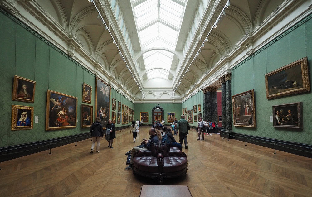 People walk and sit in an art gallery with high ceilings and paintings on green walls. The wooden floor gleams under the natural light.