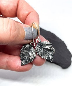 Hand holding two silver leaf-shaped earrings with a small crescent moon detail on each, against a blurred background.