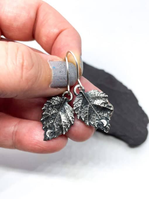 Hand holding two silver leaf-shaped earrings with a small crescent moon detail on each, against a blurred background.