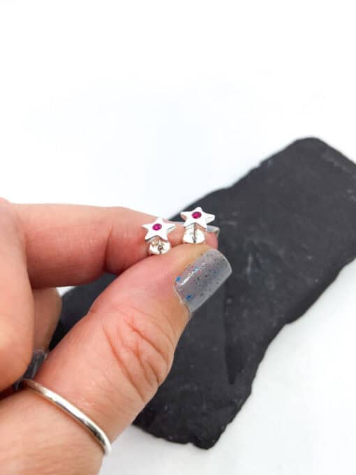 Fingers holding star-shaped silver earrings with pink gemstones against a dark stone background and white surface.