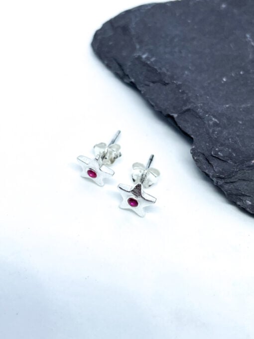 Silver star-shaped stud earrings with red center stones on a white surface, next to a piece of black slate.