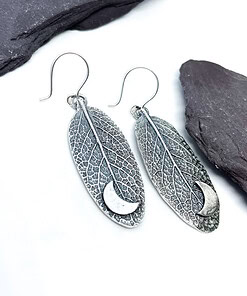 Silver leaf-shaped earrings with crescent moon cutouts, placed on a white background with dark stones.