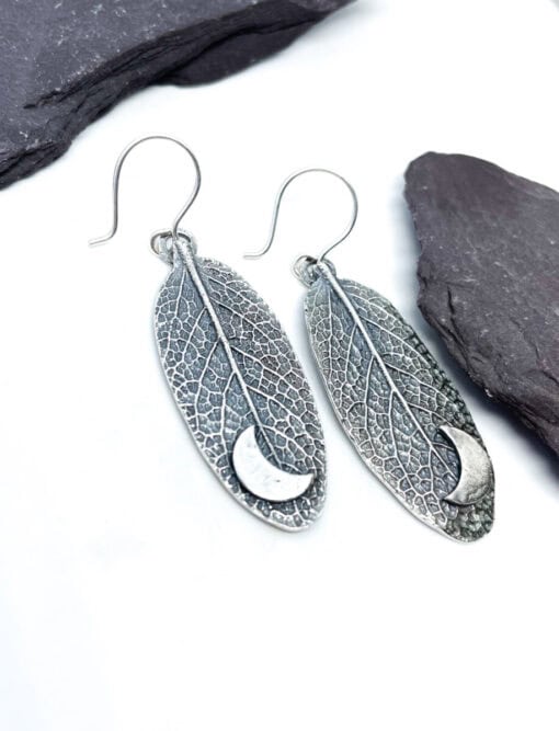 Silver leaf-shaped earrings with crescent moon cutouts, placed on a white background with dark stones.