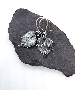 Handcrafted silver leaf-shaped earrings with intricate detailing, displayed on a flat black stone surface.