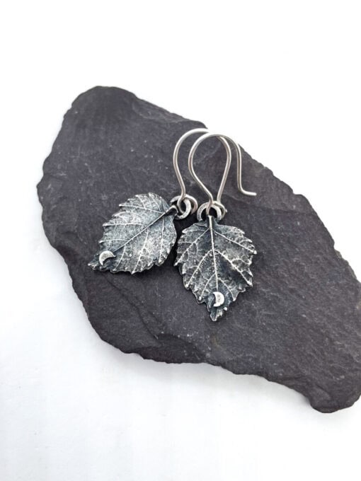 Handcrafted silver leaf-shaped earrings with intricate detailing, displayed on a flat black stone surface.