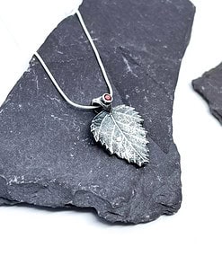 A silver leaf pendant with a small red gemstone on a thin chain, displayed on dark slate stones.