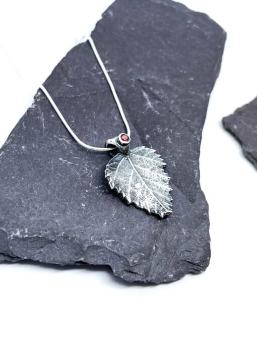 A silver leaf pendant with a small red gemstone on a thin chain, displayed on dark slate stones.