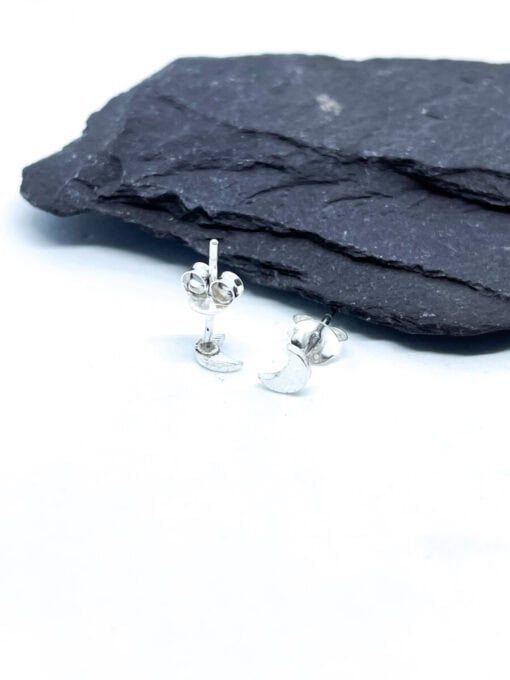 Small silver crescent moon stud earrings displayed on a textured black rock surface.