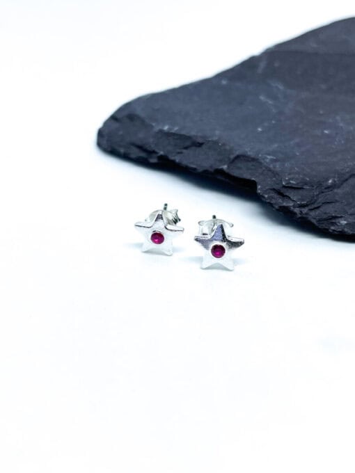 Silver star-shaped earrings with small pink gemstones in the center, placed on a white surface near a black slate.