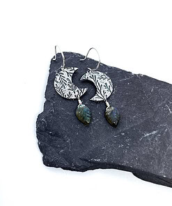 Silver crescent earrings with intricate leaf patterns and dangling green leaves, displayed on a dark slate surface.