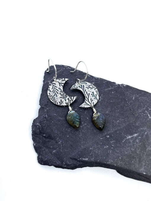 Silver crescent earrings with intricate leaf patterns and dangling green leaves, displayed on a dark slate surface.