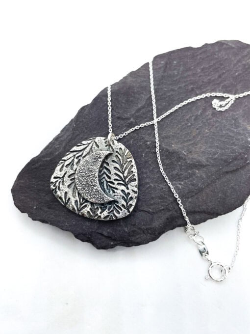 Silver necklace with a crescent moon and leaf pattern pendant, displayed on a black slate background.