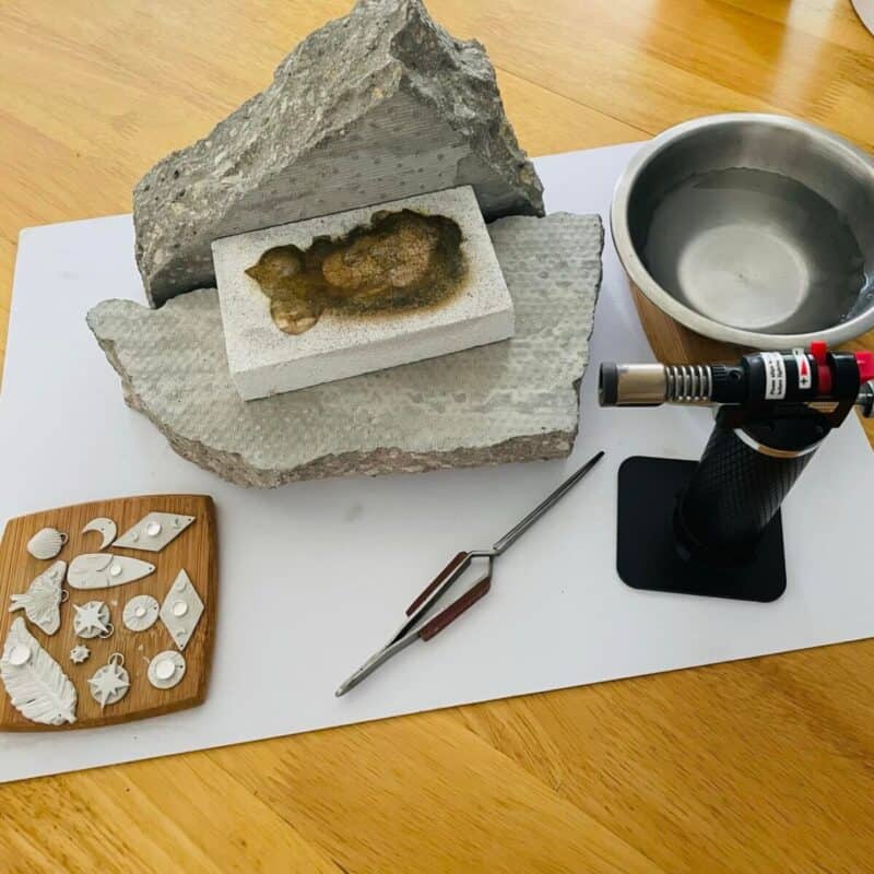 Tools for silver clay making including a blow torch, tongs, water bowl, textured rocks, and shaped silver clay pieces.