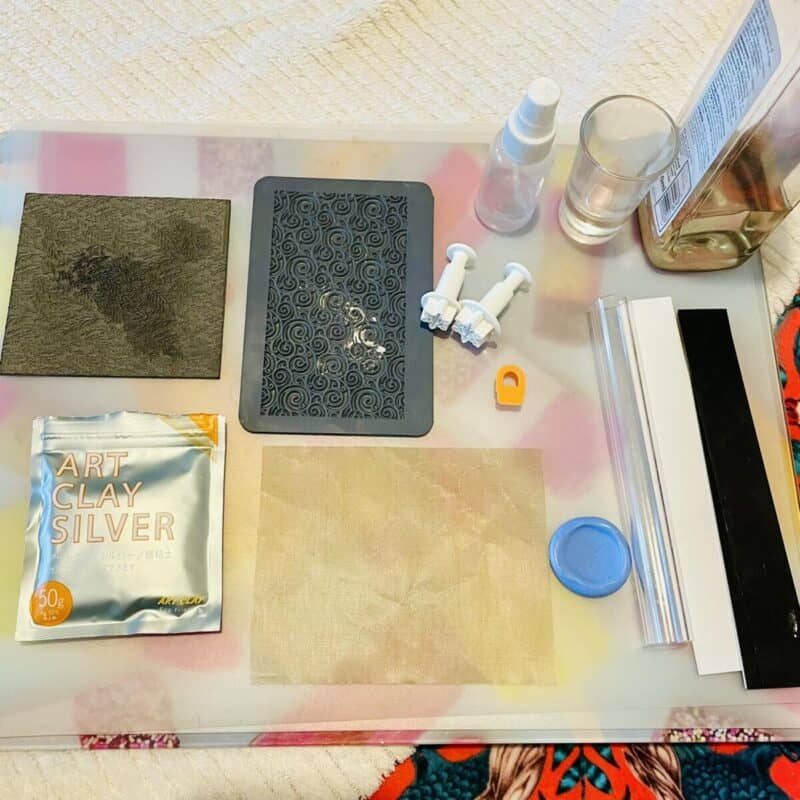 Behind the scenes of jewellery making: tools, Art Clay Silver package, molds, texture sheets, and spray bottle on a white surface.