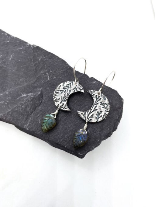 Oxidized silver earrings with floral patterns and leaf-shaped labradorite dangles, displayed on a dark stone surface.