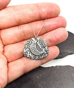 A hand holding a silver pendant with a crescent moon and leaf design, on a silver chain, against a dark background.