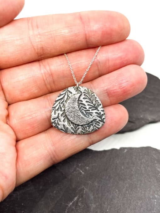 A hand holding a silver pendant with a crescent moon and leaf design, on a silver chain, against a dark background.