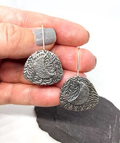 Hand holding textured metal earrings with a crescent moon and leaf design, displayed above a dark stone background.