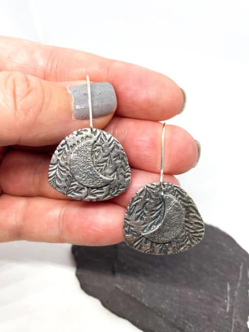 Hand holding textured metal earrings with a crescent moon and leaf design, displayed above a dark stone background.