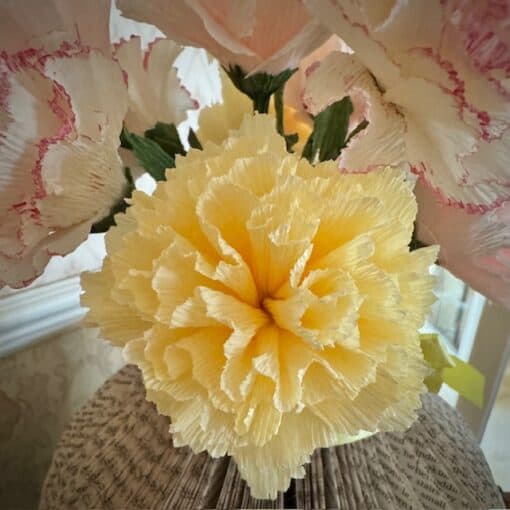 Yellow crepe paper flower surrounded by pink and white flowers, set on a background of folded book pages.