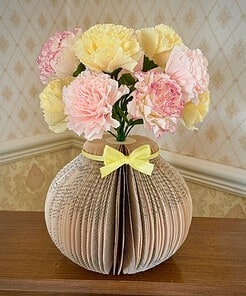 Paper flowers in a vase made from folded book pages, placed on a wooden table against a patterned wallpaper.